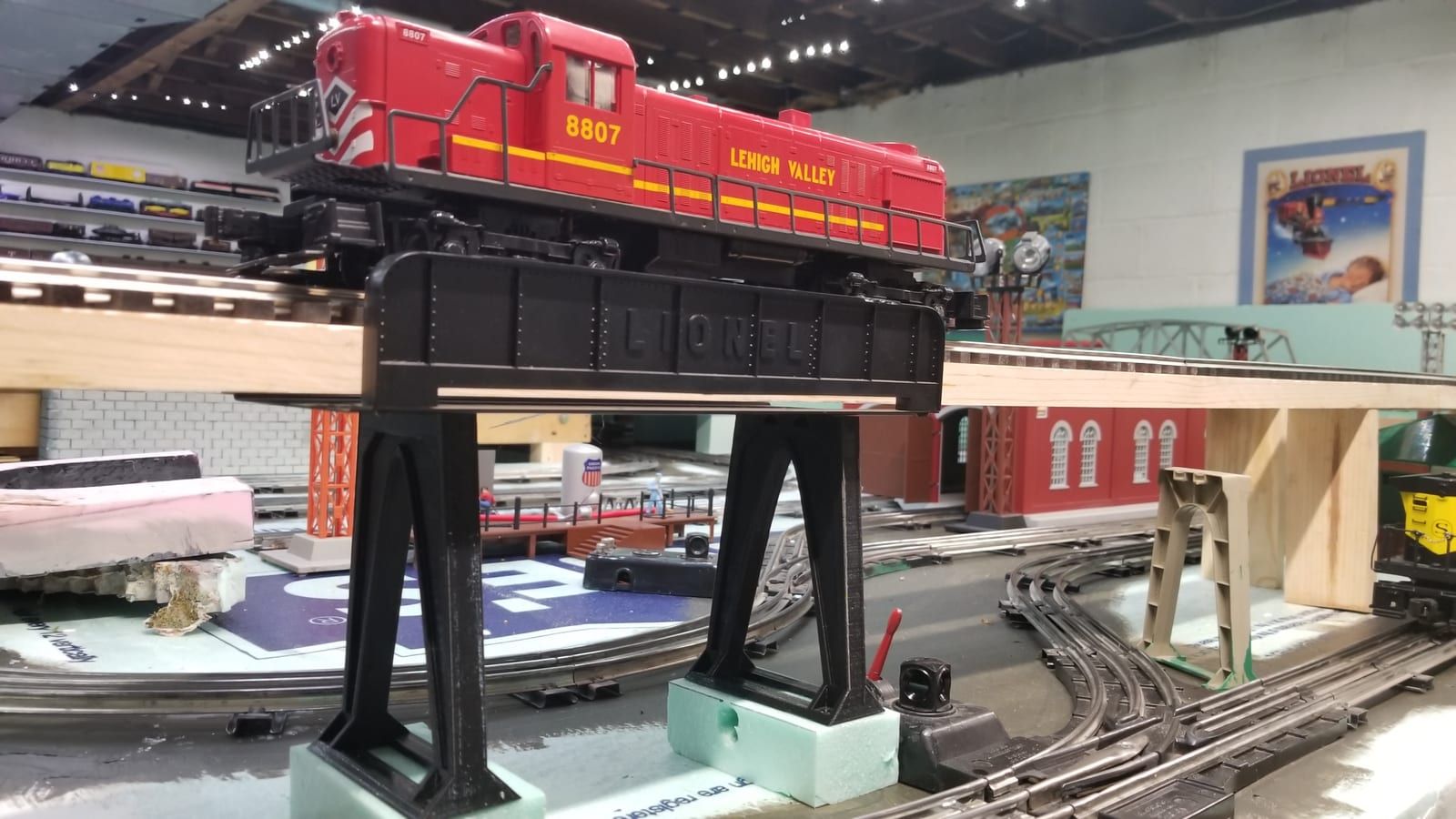 A black 3D-printed truss bridge carrying a red O-scale locomotive on a model railroad layout.